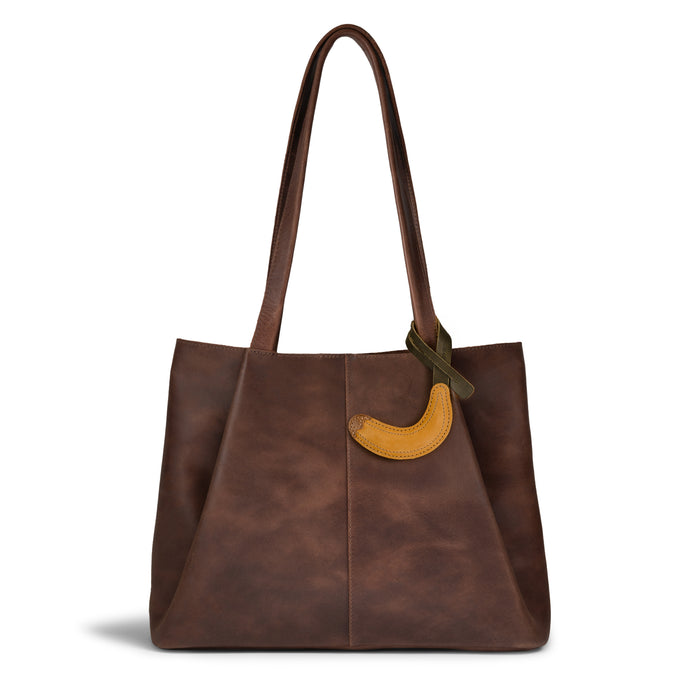 Hide & Drink Banana Bag Charm attached to a stylish brown tote bag. This accessory elevates your look and is ideal as a purse charm for totes and backpacks. Crafted from full grain leather, it's truly an ornament for women that reflects handmade quality from La Antigua Guatemala and Oaxaca Mexico, emphasizing small batch production and artisan craftsmanship.