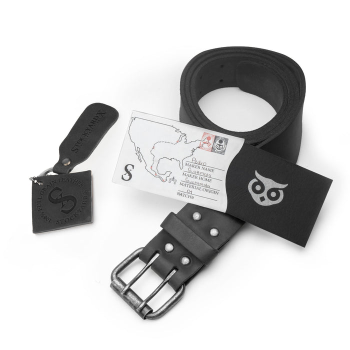 Showcasing the black Reinforced Double Prong Buckle Belt by Hide & Drink with its packaging, emphasizing its rugged design. This belt measures 1.5 in. wide and is made from full grain leather, showcasing stockyardX craftsmanship through small batch production, with details on its hand cut, hand sewn, and fire branded artisan methods, made in La Antigua Guatemala & Oaxaca Mexico.