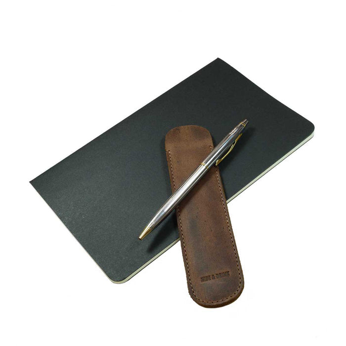 The Vintage Pen Sleeve from Hide & Drink rests alongside a notebook, emphasizing its stylish and functional design as an essential accessory for any bookworm. Made from high-quality Full Grain Leather, each piece is hand cut and hand sewn.